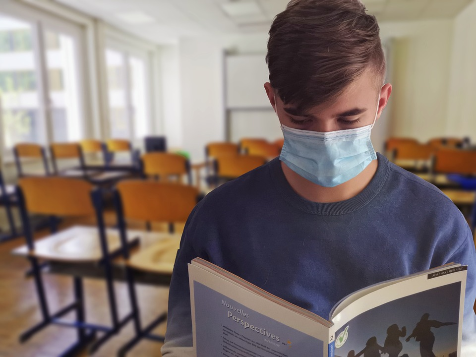 A teenage boy wearing a mask is reading a book in a classroom